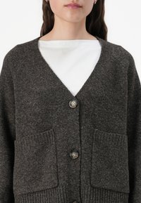 Mörkbrun ullkofta med V-hals, två framfickor och knappar med ett naturligt mönster samt textil med struktur.
