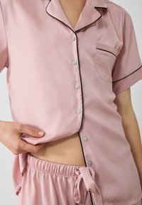 Roze satijnen pyjamasset met een kortmouwige button-up top met zwarte piping en een borstzak, gecombineerd met shorts met elastische tailleband.