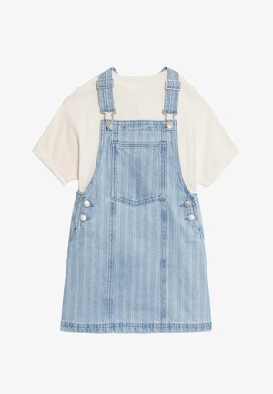 Jeans-Overallkleid mit hellblauen Streifen, kurzen Ärmeln und Knöpfen an den Taschen, kombiniert mit beigen Slip-on Schuhen. Kind in verspielter Haltung.