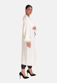 Langer weißer Pullover mit einem Kabelstrickmuster, weiten Ärmeln und einem Kordelzug in der Taille, kombiniert mit schwarzen Lederhosen und hohen Schuhen.
