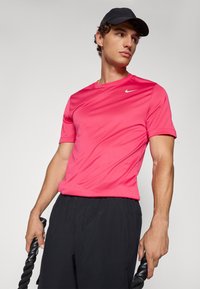 Różowa koszulka sportowa uszyta z lekkiego, gładkiego materiału, z okrągłym dekoltem i małym białym logo. W zestawie z czarnymi szortami i trzymającą linę.