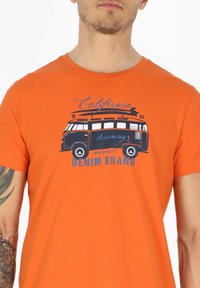 Oranssi puuvillapaita, jossa on vintage-bussin grafiikka ja "California dreaming" -teksti sinisellä, pyöreä kaula-aukko ja lyhyet hihat.