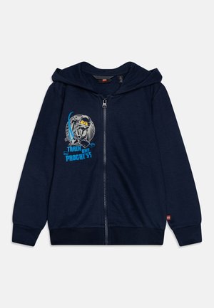 Sweat à capuche zippé bleu marine avec capuche, affichant un graphique de ninja LEGO et le texte "Entraîne-toi et progresse" en bleu sur le côté gauche de la poitrine.
