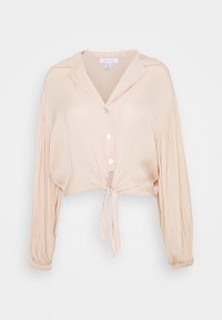 Kermanvärinen lyhyt paita, jossa on pitkät hihat, napitettava etuosa ja nauhamainen yksityiskohta vyötäröllä, yksinkertaista valkoista taustaa vasten.