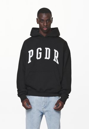 Mladý muž v čiernej mikine s kapucňou, na ktorej sú biele písmená "PGDR", a v svetlomodrých džínsoch, stojaci na bielom pozadí.