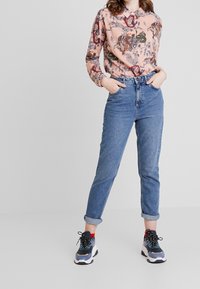 Langærmet ferskenfarvet skjorte med flerfarvede blomster- og dyreprint, parret med højtaljede, oprullede blå jeans og sorte atletiske sneakers.