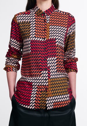 Donna che indossa una camicia a maniche lunghe con bottoni e motivo geometrico in rosso, arancione, marrone e crema, mani nelle tasche dei pantaloni neri.