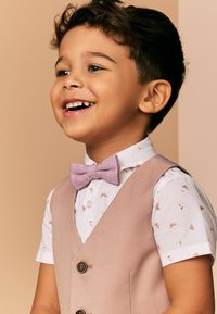 Gilet rose clair sur une chemise blanche à motifs floraux, avec un nœud papillon violet. Le garçon a les cheveux courts et bouclés et arbore une expression souriante.