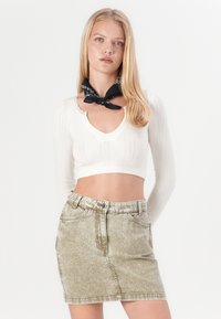 Fehér, bordázott crop top V-nyakkal és hosszú ujjakkal, egy elhalványult olívazöld farmer miniszoknyával párosítva. Fekete sál, a nyaknál megkötve.