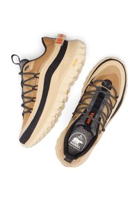 Chaussures de sport beige et noires avec matériaux texturés, semelles épaisses blanches et lacets noirs. Présentent des accents orange et le logo Sorel.