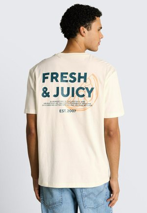 Mężczyzna zwrócony bokiem, ubrany w kremowy t-shirt z napisem „FRESH & JUICY” i grafiką plastra pomarańczy na plecach, w połączeniu z niebieskimi dżinsami.