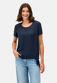 Blouse courte à manches courtes bleu marine en tissu lisse, dotée d'un col rond et d'un détail de nouage sur le côté pour un intérêt supplémentaire. Associée à un jean clair.