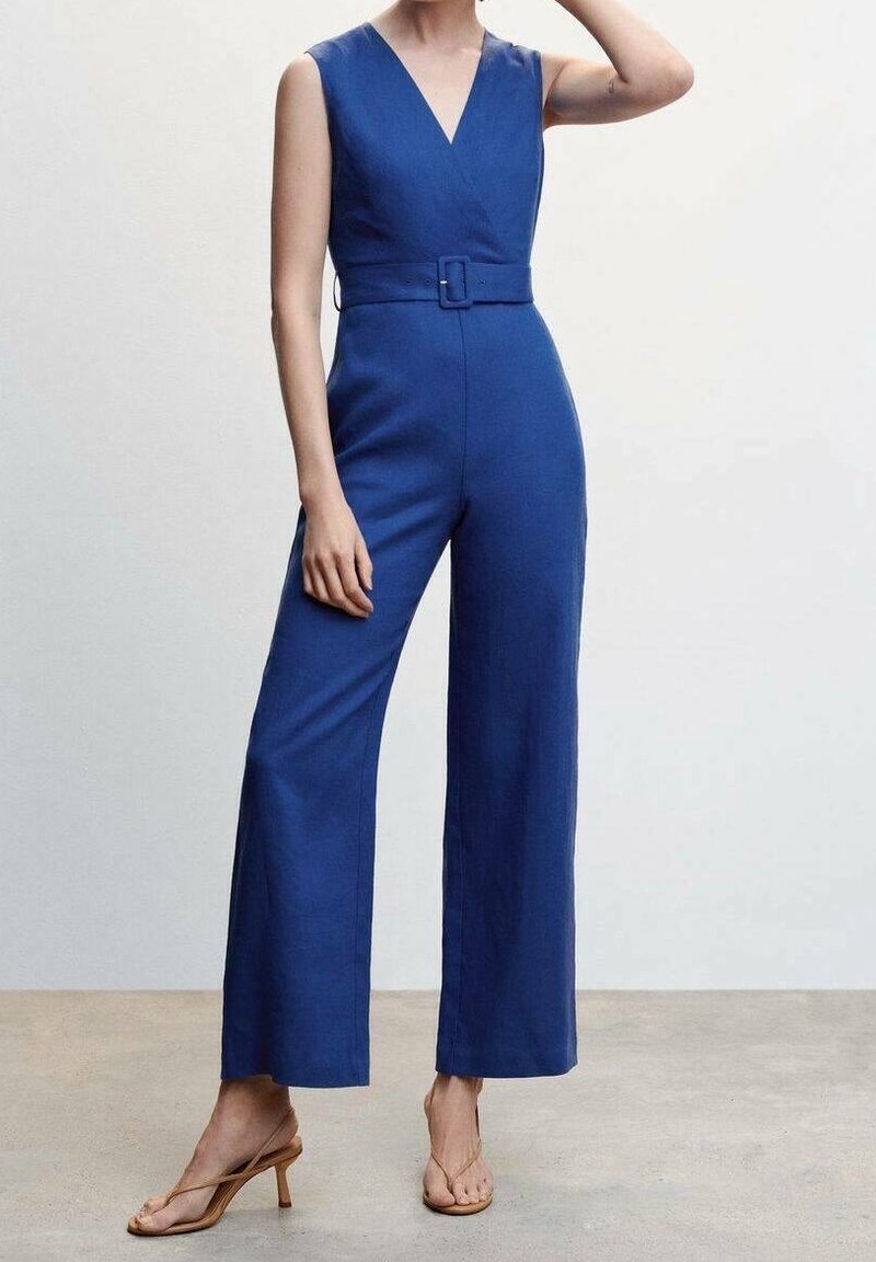 Femme portant une combinaison bleu sans manches à jambes larges avec une ceinture assortie, associée à des sandales à talons hauts couleur nude sur un fond uni.