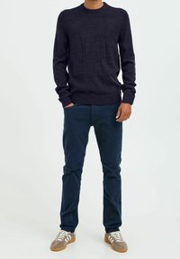Mann trägt einen dunkelblauen Pullover, dunkle blaue Hosen und beige Turnschuhe mit weißen Streifen, steht mit einer Hand in der Tasche vor einem weißen Hintergrund.