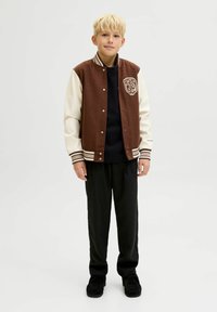 Pruun ja kreemi värvi ülikooli jakki koos ribatud varrukate ja kraega, millel on esiküljel logo; kombineeritud mustade pükste ja tumedate kingadega.