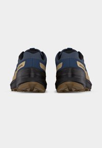 Ζεύγος παπουτσιών τρεξίματος στη φύση Salomon σε ναυτικό μπλε και ταμπέ χρώμα, εμφανιζόμενα από πίσω σε απλό φωτεινό γκρι φόντο.