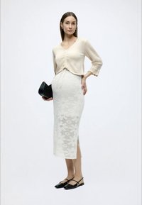 Εκρού πλεκτή κοντή ζακέτα με τελείωμα δαντέλας, συνδυασμένη με λευκή, πλεκτή midi φούστα με λουλουδένιο μοτίβο. Μαύρη τσάντα και επίπεδα παπούτσια.