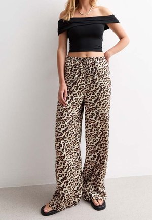 Nainen, jolla on musta olkapäät paljastava lyhyt toppi, väljähköt leopardi-kuviolliset housut nyörillä ja mustat varvassandaalit, seisoo yksivärisen valkoisen seinän edessä.