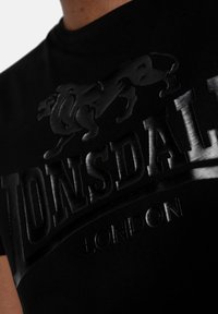 Camiseta negra con un diseño gráfico en relieve de un león y el nombre de la marca "LONSDALE LONDON" en acabado brillante en la parte frontal.