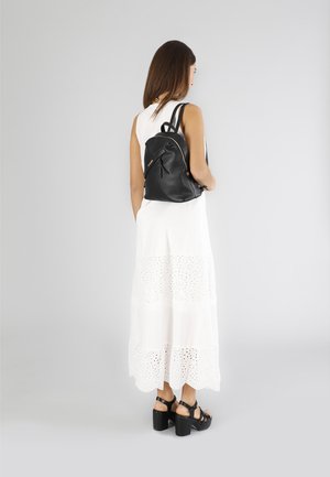 Sac à dos en cuir noir avec une finition texturée, des détails en zip et des bretelles ajustables, porté sur une robe blanche à volants en dentelle.