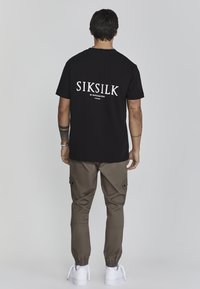 Black cotton t-shirt with "SIKSILK" logo in white on the back, paired with olive green jogger pants and white sneakers.