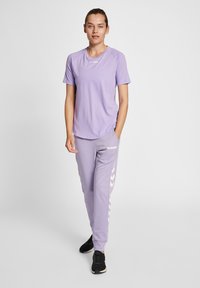 Lavendel atletisk t-shirt och matchande joggers med vita chevronränder. Materialien verkar lätta. Modellen bär svarta sneakers.