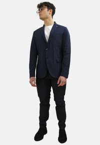 Blazer navy sartoriale con un solo bottone, rever a scendenza e tasche laterali, abbinato a pantaloni neri a gamba affusolata e scarpe scure.