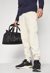 Jogging pants crème avec des poignets élastiques, portés avec un sweat-shirt bleu marine, des chaussures de sport noires, et tenant un sac de sport noir avec des accents blancs.