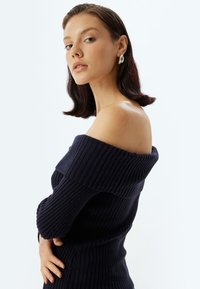 Schulterfreier, gerippter Pullover in Dunkelblau. Strukturierter Strick mit langen Ärmeln und einer figurbetonten Silhouette. Modell posiert mit der Hand auf der Hüfte.