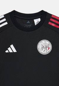 Zwart voetbalshirt met ronde hals, voorzien van witte Adidas-strepen op de schouders en een cirkelvormig Ajax-logo met een gedetailleerde illustratie.