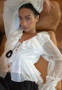 Blusa bianca a volant con maniche lunghe e trasparenti, dettagli con cordoncino, indossata sopra pantaloni neri; accessoriate con una grande collana con pendente scuro.