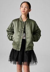 Giacca bomber in satin verde oliva con colletto e polsini a coste, dotata di tasche laterali e chiusura con zip, indossata sopra una gonna in tulle nera.
