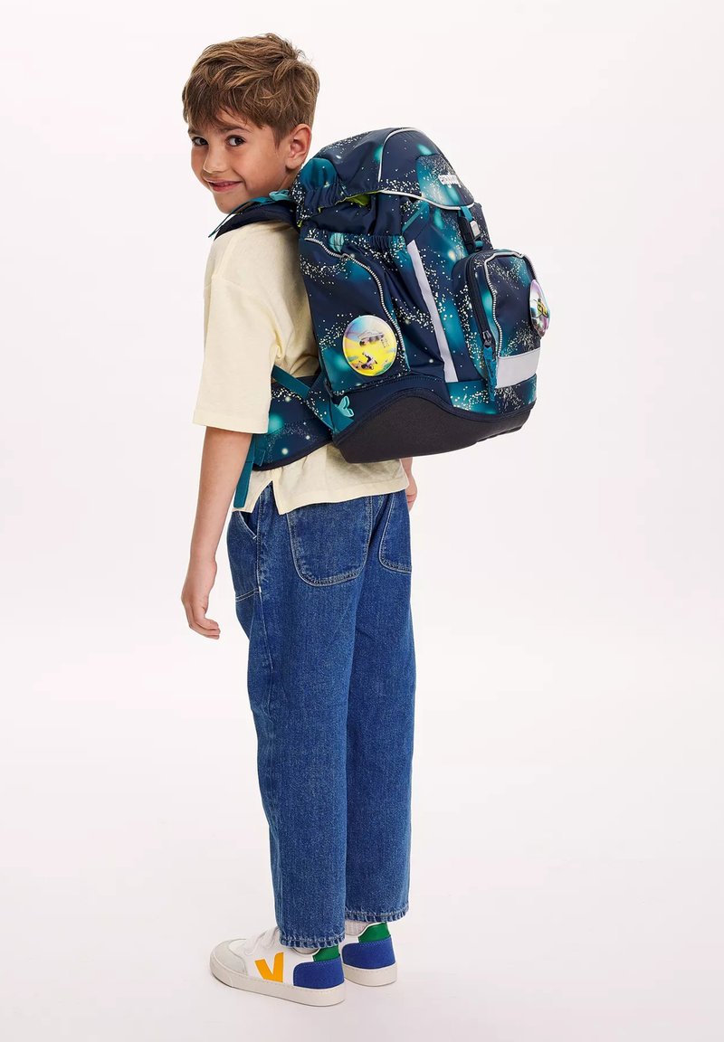 Junge lächelt über die Schulter, trägt ein gelbes Hemd, blaue Jeans, weiße Turnschuhe und einen großen Rucksack mit Galaxie-Muster und bunten Aufnähern.