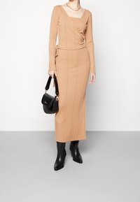 Robe longue à côtes en beige clair avec un design croisé, des manches longues et une fente jusqu'à la cuisse. Associée à des bottes en cuir noir jusqu'au genou et un sac à main.