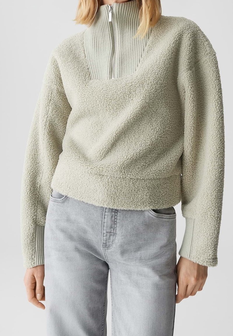Femme portant un pull sherpa polaire gris clair avec col zippé côtelé et jean gris clair, debout devant un fond uni.