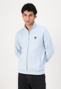 Sweatshirt azul claro com fecho zip, feito de tecido macio, apresentando um alto colar, bolso lateral e um distintivo de logo no peito esquerdo.