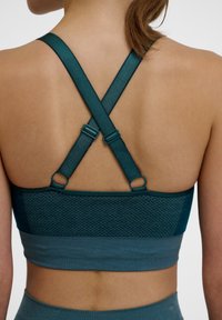 Teal sportbeha met kruisbanden achter met verstelbare bevestigingen, een geperforeerde mesh rugpaneel en een gladde band langs de onderrand.