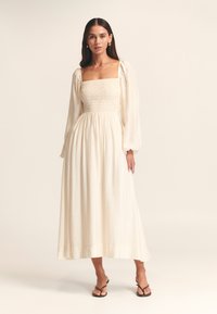 Lichtbeige jurk met een gesmokt lijfje, lange pofmouwen en een vloeiende rok. Gemaakt van een textiel met structuur, enkellange silhouet.