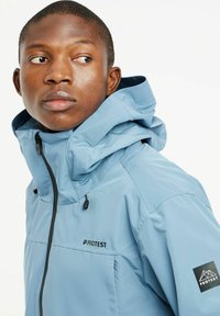 Šviesiai mėlynas gobtuvas striukė su užtrauktuku, papuošta tamsiomis siūlėmis ir logotipais ant krūtinės ir rankovės. Sklandi, vandeniui atspari medžiaga.