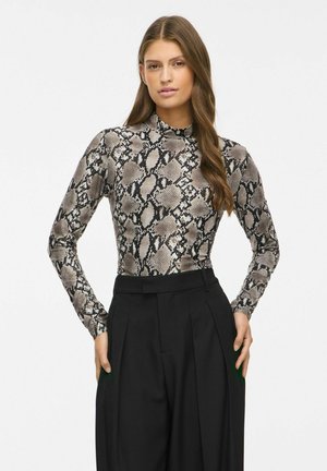 Femme aux longs cheveux bruns portant un haut à col montant à manches longues imprimé serpent et un pantalon noir taille haute à jambes larges, debout devant un fond uni.