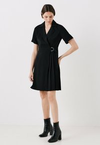 Robe noire à manches courtes en wrap avec une taille ceinturée et une jupe plissée. Tissu lisse et col pointu ; portée avec des bottines à talons noires.