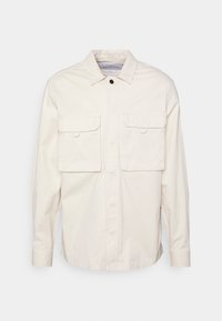 Jasnobeżowa koszula na guziki wykonana z bawełny, z dwoma kieszeniami na piersi z klapami i klasycznym kołnierzem. Prosty design z minimalnymi zdobieniami.