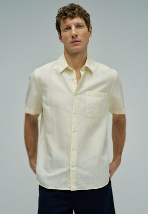 Hombre con pelo rizado que lleva una camisa de manga corta color crema con botones, sin meter por dentro, y pantalones oscuros, de pie con las manos detrás de la espalda.