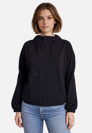 Jonge vrouw draagt een zwarte hoodie met rits, trekkoorden en elastische boorden, gecombineerd met lichtblauwe spijkerbroek, staand tegen een witte achtergrond.