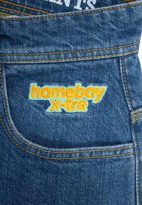 Denim jeans med blått stoff og oransje sømmer, samt en lys brodert logo med teksten "homeboy x-tra" på baklommen.