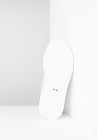 Bílá podrážka tenisky s texturovaným vzorem a malou italskou vlajkou, umístěná vzpřímeně na pozadí v bílé a šedé barvě.