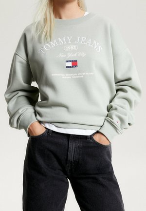 Persona que lleva una sudadera gris claro de Tommy Jeans con texto New York City, logo y vaqueros negros, manos en los bolsillos, mostrada desde el cuello hasta las caderas.
