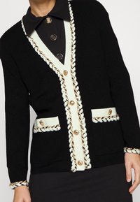 Cárdigan negro con detalles crema, tejido texturizado, botones dorados y ribete trenzado en los bordes y bolsillos. Ajuste entallado, cuello clásico.