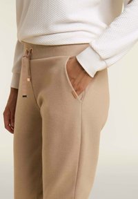 Pantalones jogger beige hechos de tela suave, con cintura ajustable con cordón, bolsillos laterales y un pequeño detalle de marca en el bolsillo.