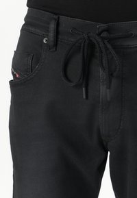 Črne hlače iz gladkega blaga z vrvico v pasu, ki imajo sprednji žep z okrasno nalepko in subtilnimi šivi.